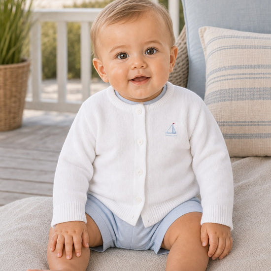 Carriage Boutique white cotton baby cardigan with sailboat embroidery, worn by a smiling baby on a porch.