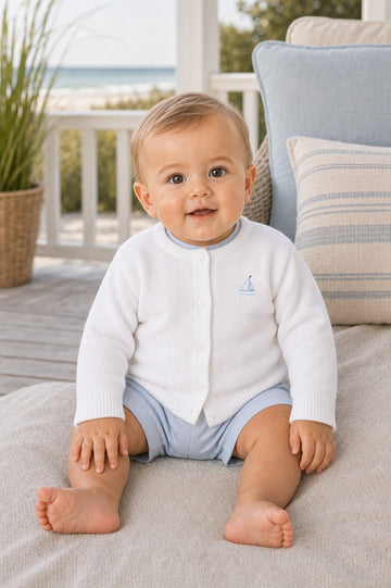 Carriage Boutique white cotton baby cardigan with sailboat embroidery, worn by a smiling baby on a porch.