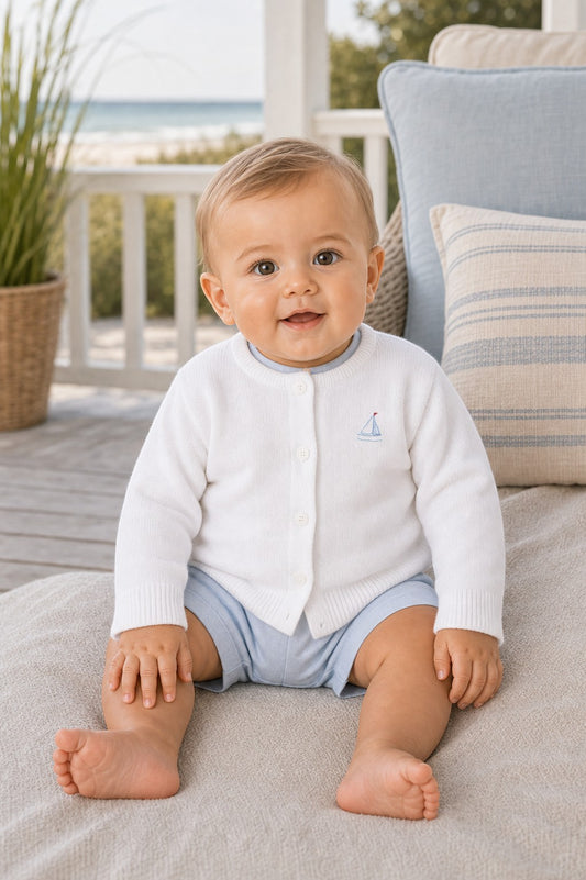 Carriage Boutique white cotton baby cardigan with sailboat embroidery, worn by a smiling baby on a porch.