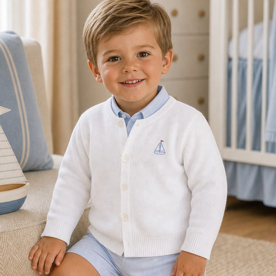 Carriage Boutique white cotton cardigan for boys featuring a small blue sailboat embroidery and button-front closure.