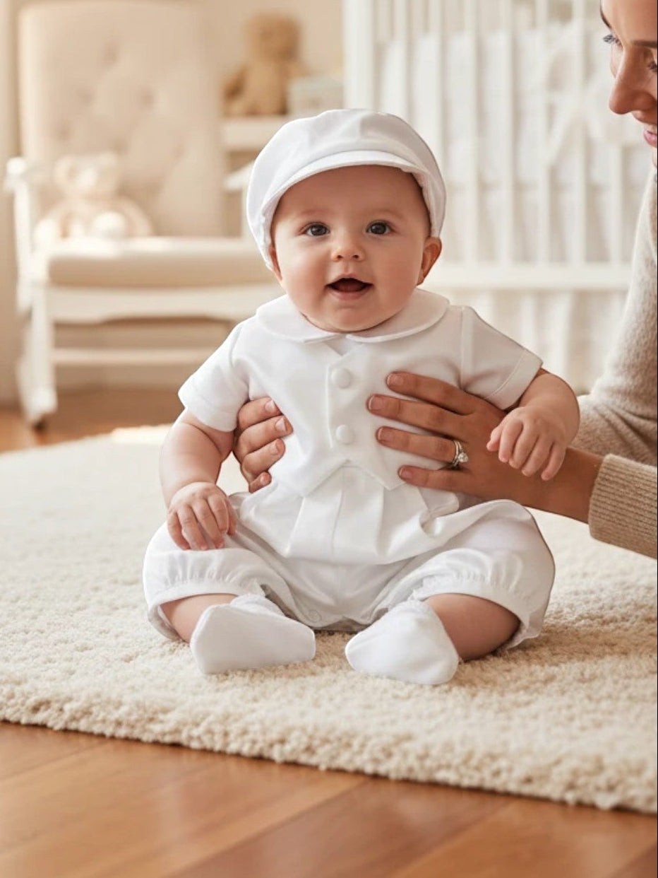 Baby boy wearing a white satin Carriage Boutique christening romper and matching cap, held by an adult.