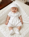 Carriage Boutique baby boy wearing a white satin christening romper with a matching hat, sitting on a lace blanket.