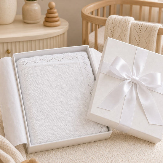 Carriage Boutique white cotton knit baby blanket with scalloped edges in a gift box on a nursery chair