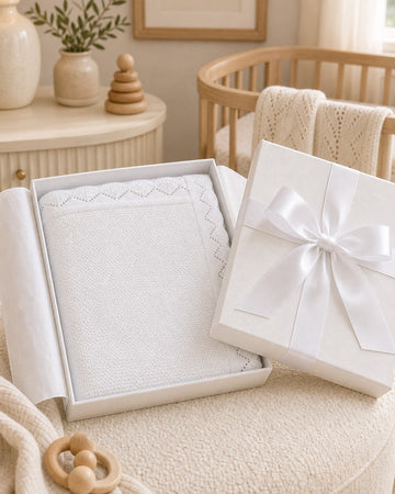Carriage Boutique white cotton knit baby blanket with scalloped edges in a gift box on a nursery chair