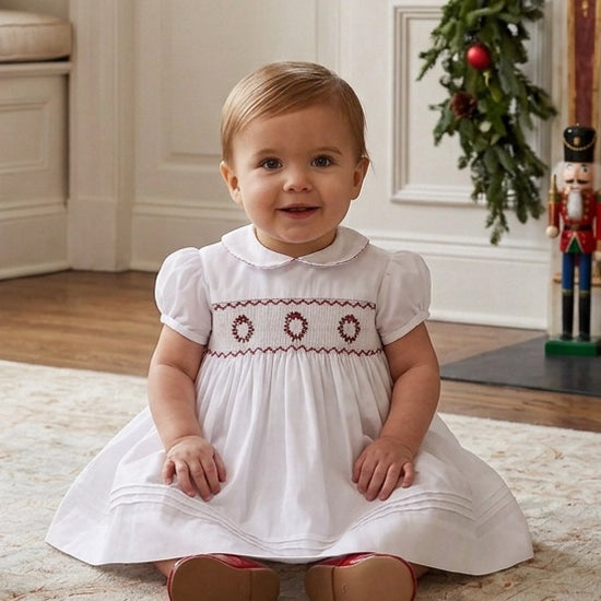 Carriage Boutique white baby dress with hand smocking and red wreath embroidery, featuring a peter pan collar.