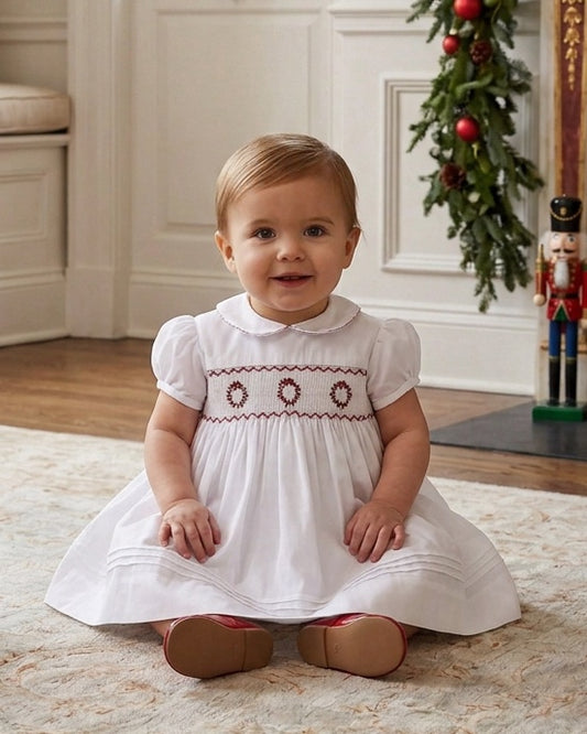 Carriage Boutique white baby dress with hand smocking and red wreath embroidery, featuring a peter pan collar.