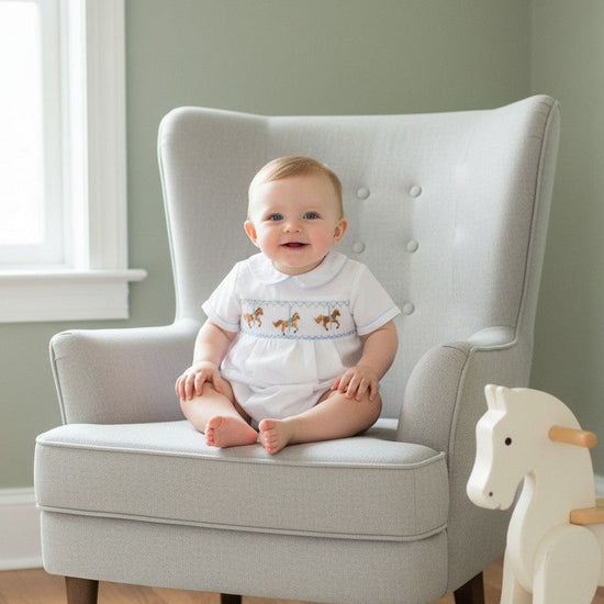 Carriage Boutique white cotton baby boy romper with hand-smocked carousel horse design and blue trim.