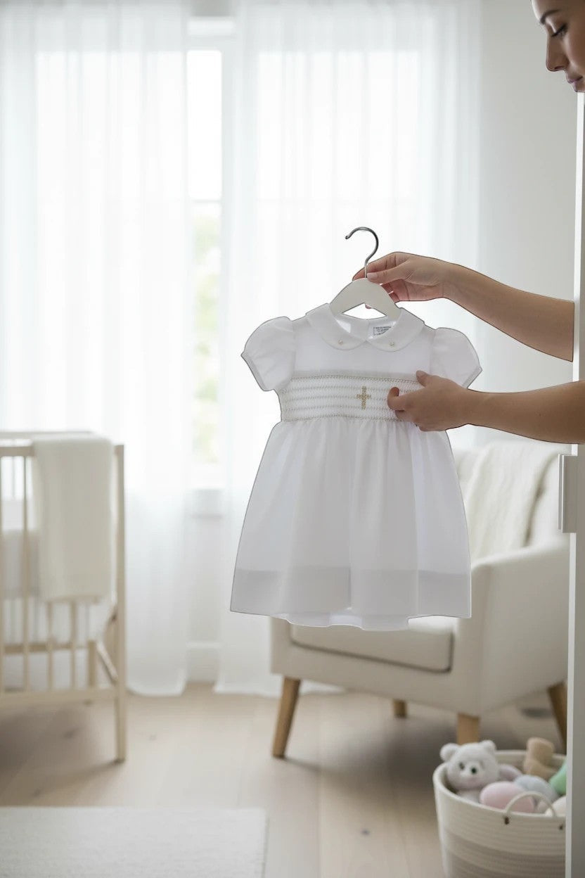 Carriage Boutique white baby girl christening dress with smocked bodice and cross embroidery in a bright nursery.