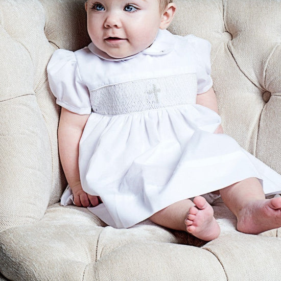 Carriage Boutique baby girl white christening dress with smocked bodice and cross embroidery sitting in a chair