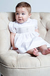 Carriage Boutique baby girl white christening dress with smocked bodice and cross embroidery sitting in a chair