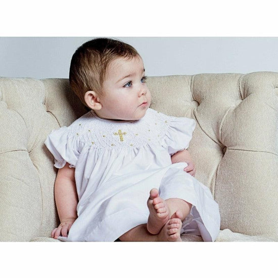 Carriage Boutique baby girl wearing a white cotton smocked bishop dress with gold cross detail and matching bonnet