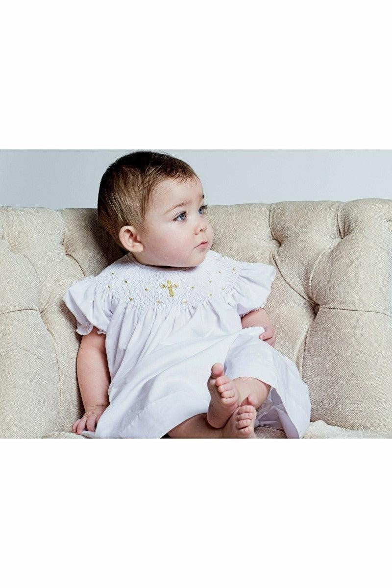 Carriage Boutique baby girl wearing a white cotton smocked bishop dress with gold cross detail and matching bonnet
