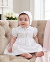 Carriage Boutique baby girl in a white smocked christening dress with a cross motif and matching bonnet.
