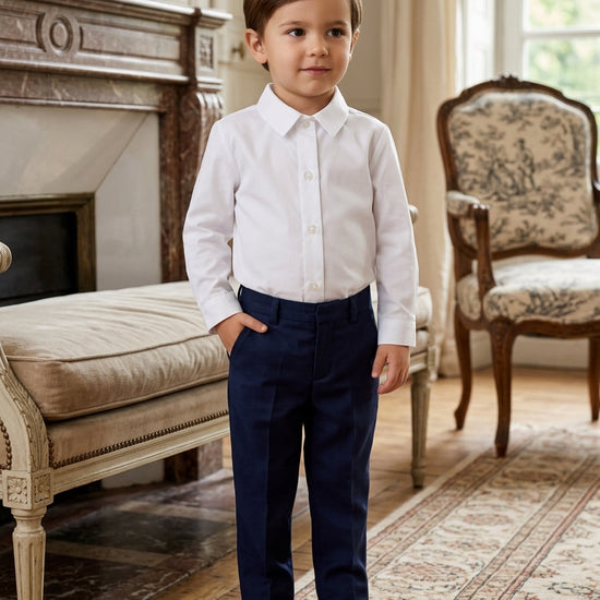 Carriage Boutique toddler boy wearing a crisp white long-sleeve button-down shirt with navy trousers and brown shoes.