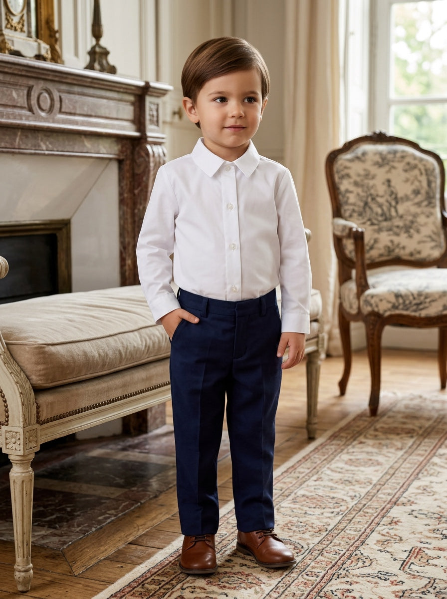 Carriage Boutique toddler boy wearing a crisp white long-sleeve button-down shirt with navy trousers and brown shoes.