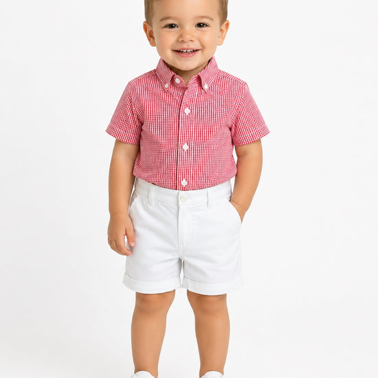 Toddler boy wearing Carriage Boutique white twill shorts and a red gingham shirt against a plain white background
