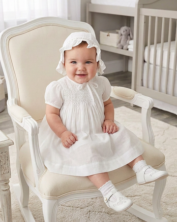 Carriage Boutique baby girl in a white voile christening dress and matching bonnet sitting on a vintage chair