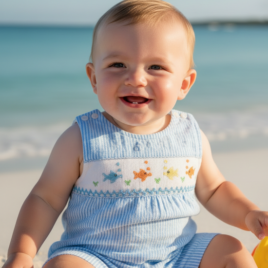Baby sitting on a sandy beach wearing a light blue outfit with colorful fish patterns.