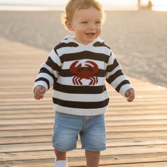 A toddler walking on a wooden boardwalk at the beach, wearing a black and white striped sweater with a red crab graphic, denim shorts, and white shoes.