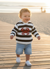 A toddler walking on a wooden boardwalk at the beach, wearing a black and white striped sweater with a red crab graphic, denim shorts, and white shoes.
