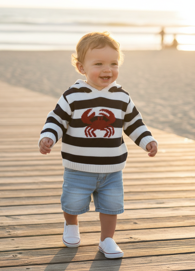 A toddler walking on a wooden boardwalk at the beach, wearing a black and white striped sweater with a red crab graphic, denim shorts, and white shoes.